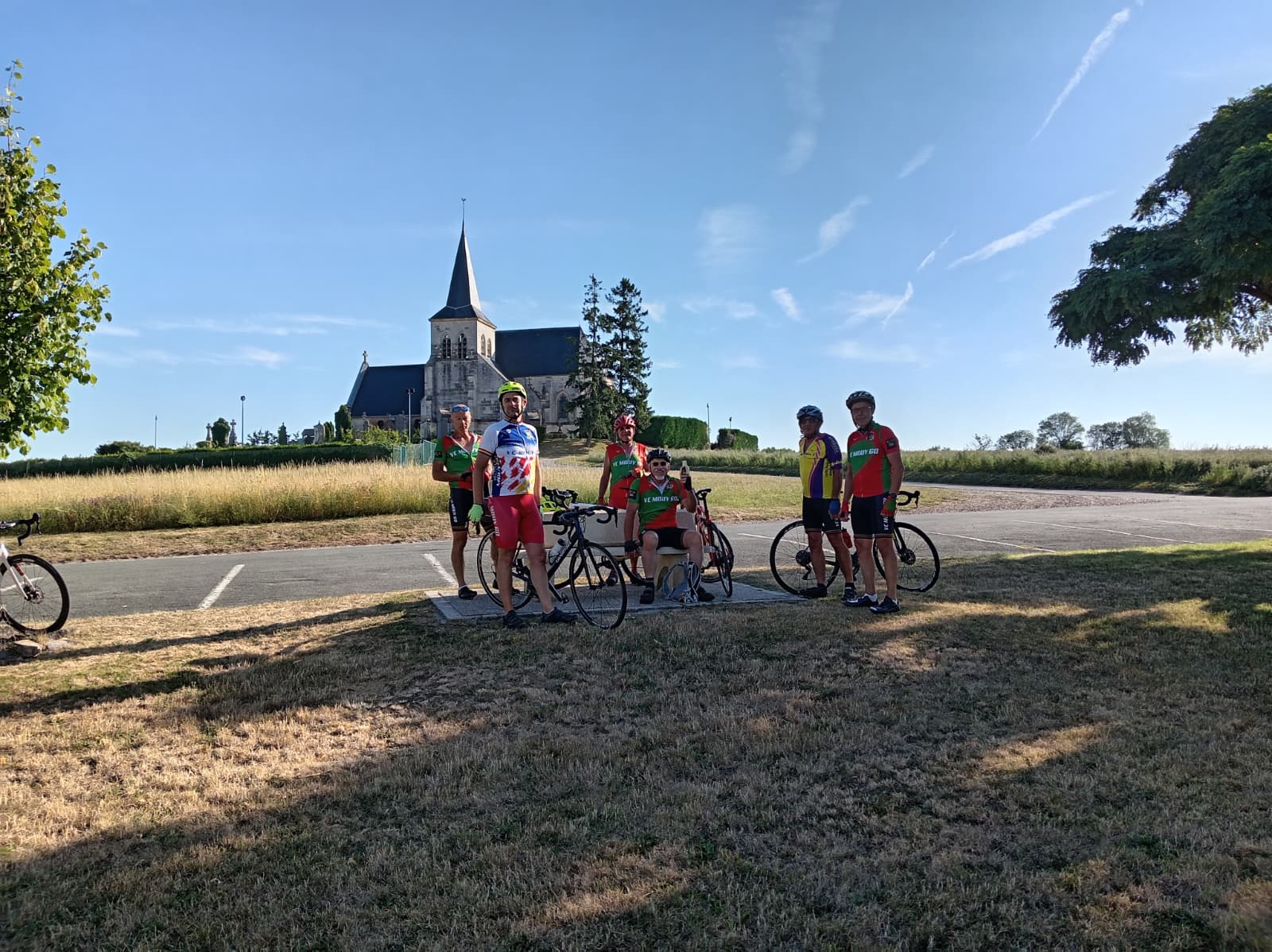 Le groupe du Vélo Club de Mouy réuni autour du banc pour la pause fraîcheur, l'église de Saint-Sulpice (Oise) en arrière-plan sous le ciel bleu
