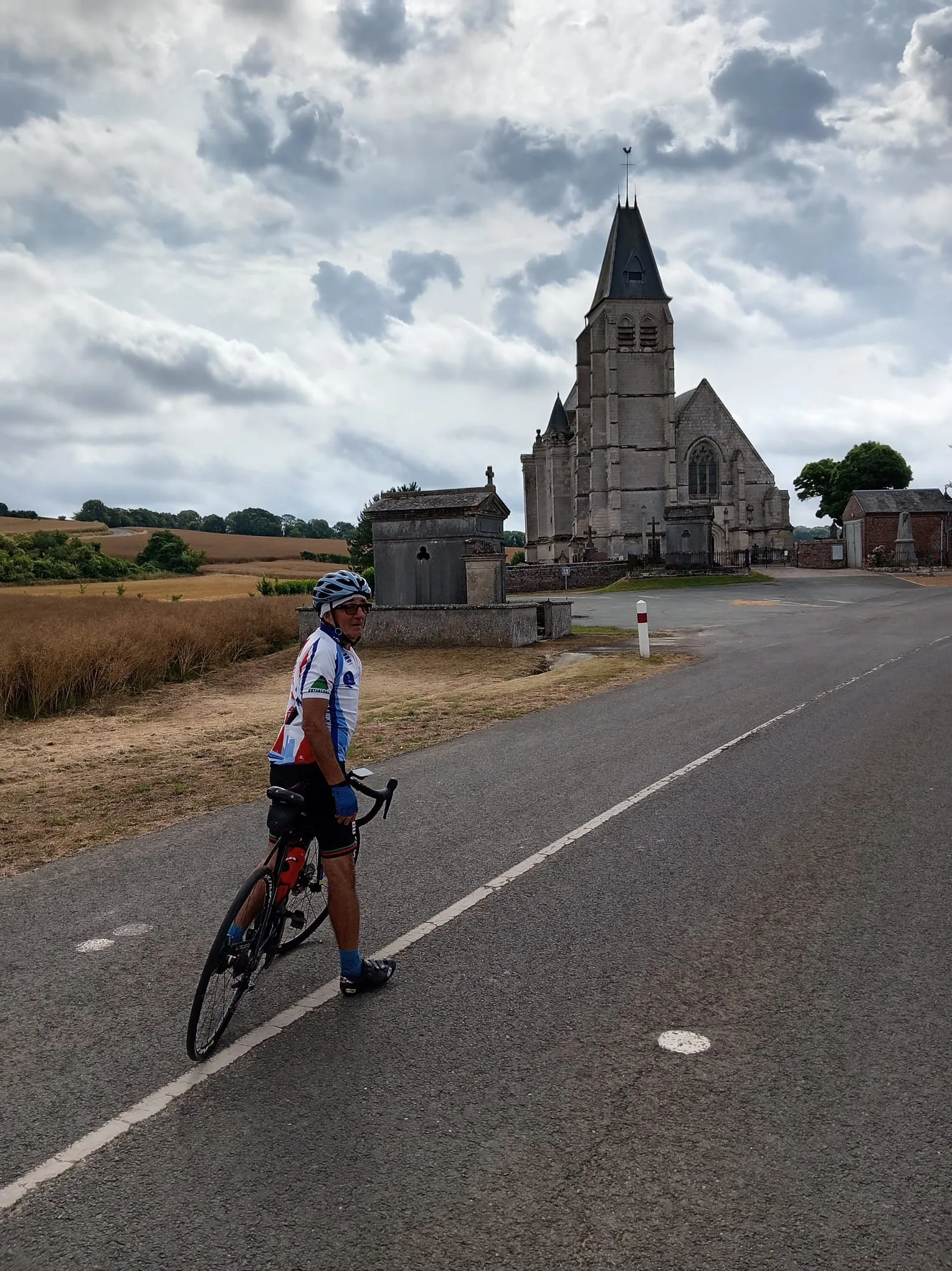 Cycliste en maillot du Vélo Club de Mouy posant devant l'église Saint-André de Saint-André-Farivillers, au milieu des champs moissonnés
