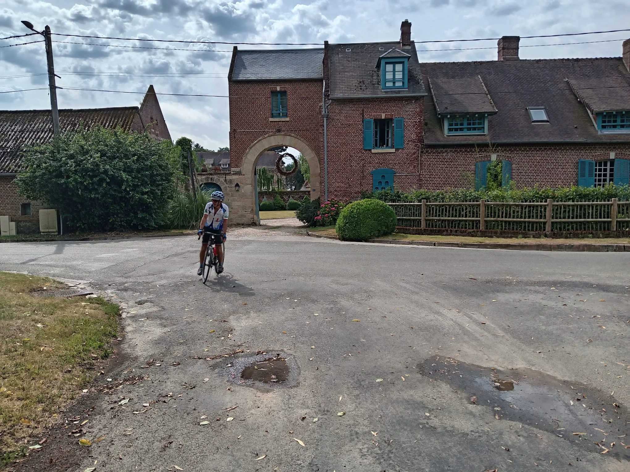 Cycliste du Vélo Club de Mouy passant devant une ferme picarde à porche en briques rouges et volets bleus, sur une petite route de campagne de l'Oise